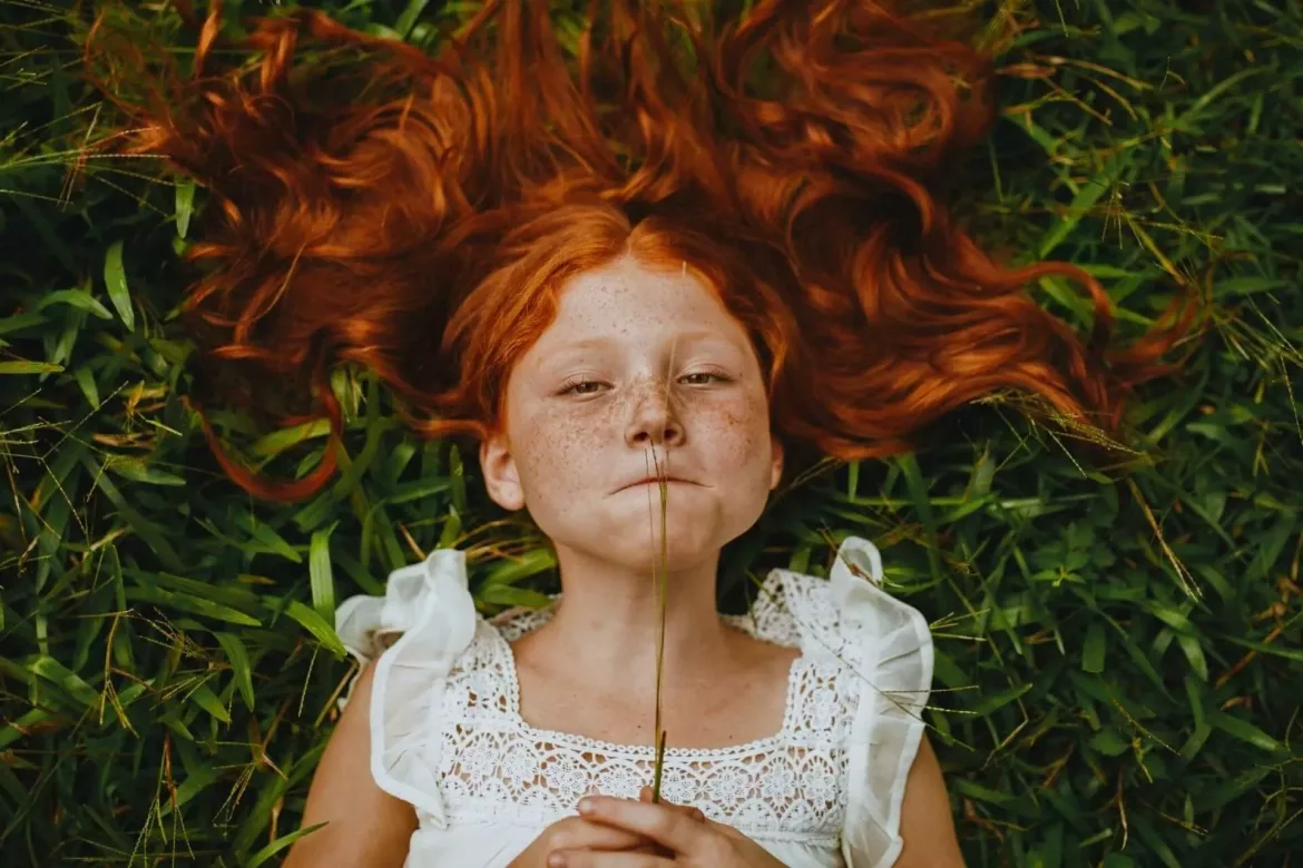 Ruda dziewczynka ze źdźbłem trawy w ustach z aparatem na zębach leży na trawie