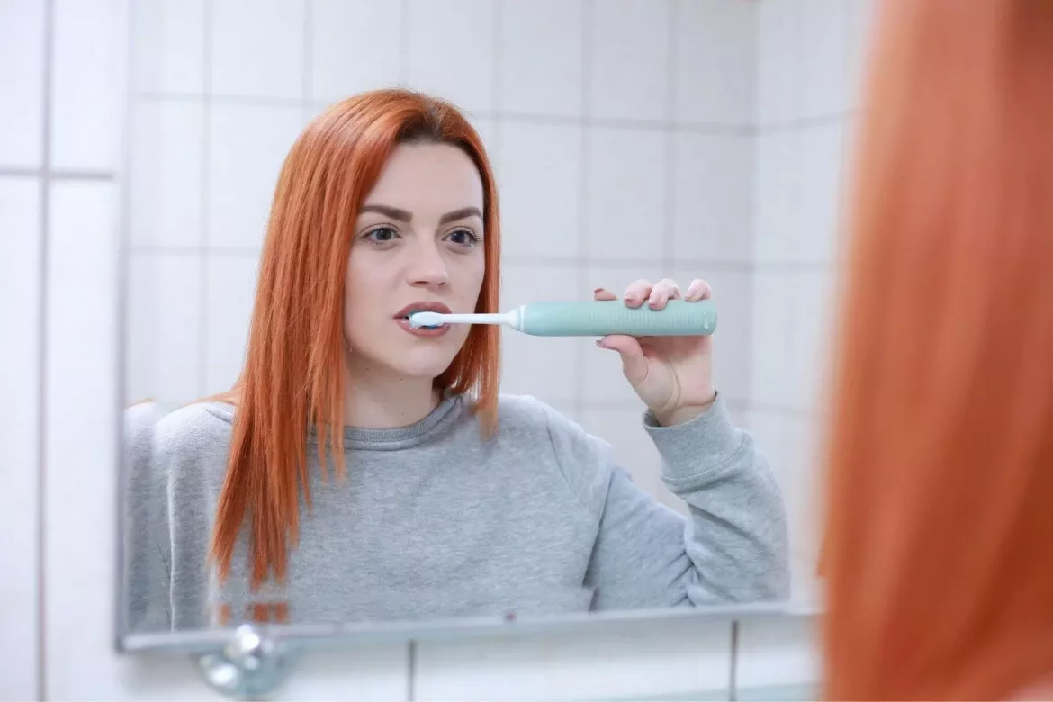 Ruda dziewczyna przed lustrem myje zęby szkoteczką elektryczną